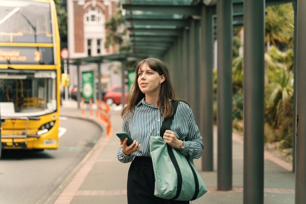 Woman looking left waiting for public bus, double decker bus in background. 