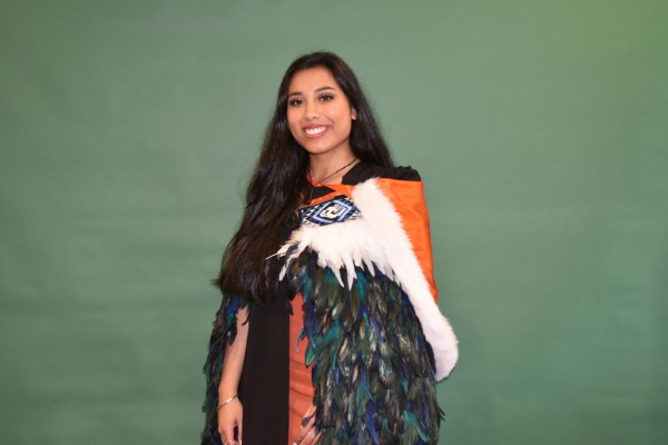 Young woman stands in front of green background, smiling, wearing an orange fur lined graduation hood with a Maaori feathered cloak.