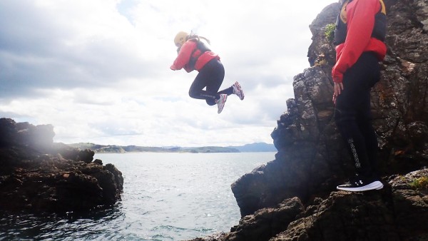 Adventure Specialties trust, girl jumps off rock into ocean watched on by another girl. Girls wear bright red jacket and black pants.