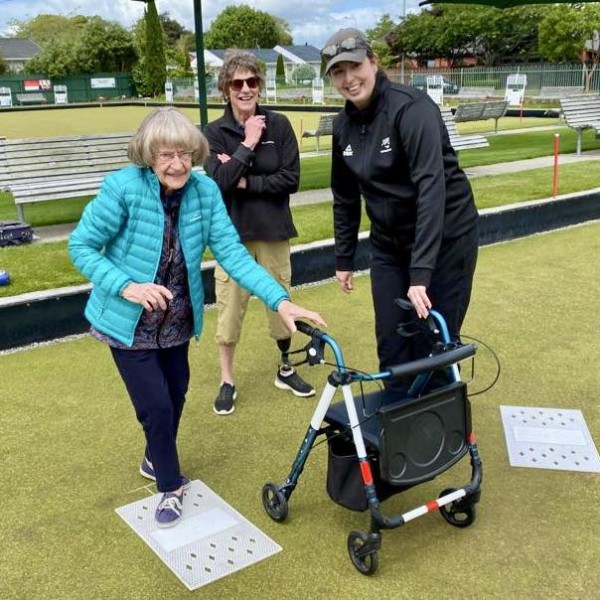 Three women play bowls. Older woman holds onto walking frame on left supported by younger woman on right. One person stands behind.