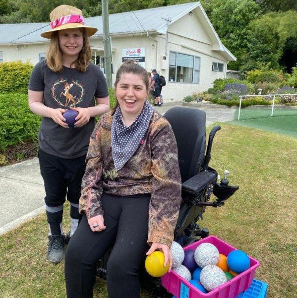 Two women smile next to box of lawn bowls. Woman in front in wheelchair. 