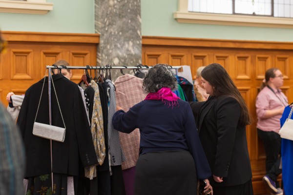 Two women look through the clothes hanging on a rack at the dress for success event. They have pulled out a pink checkered blazer and are looking at it.