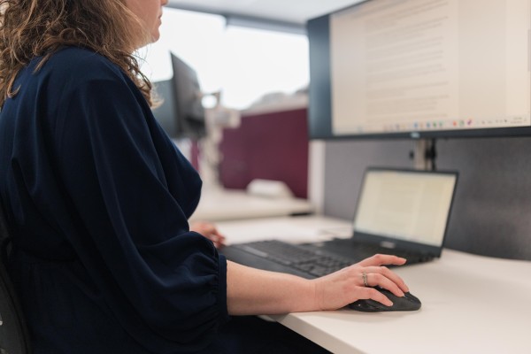 Woman in dark blue dress sits at a desk holding a computer mouse. She has a finger raised ready to click. There is a big computer screen and a laptop in front of her. She is reading words on the big screen.