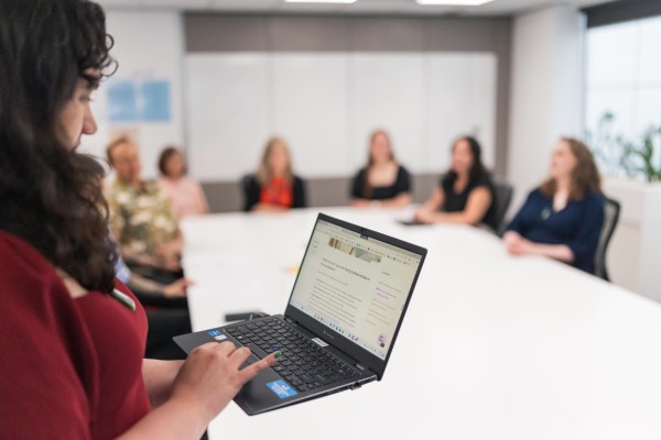 A woman with curly hair and wearing a red dress stands in front of a room of six other people with her computer. She is reading off the screen. The people in the background are blurry.