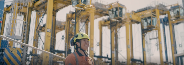 A young woman stands outside underneath large yellow cranes. She is smiling and looking into the distance on the right. She is wearing a yellow hard hat and a hi-vis jacket.