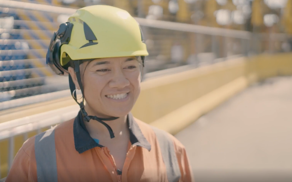 Woman wears a yellow hard hat and an orange hi-vis jacket. She is smiling at the camera. She is standing outside in an industrial area.