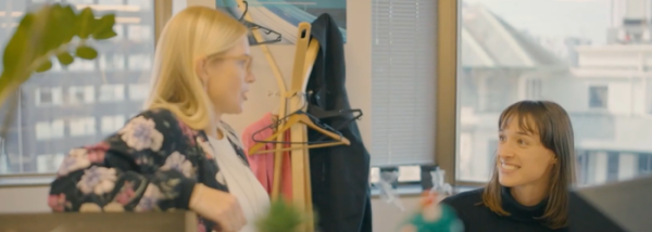A blond woman stands side on with her arm resting on a cabinet while talking to a younger woman sitting at her desk