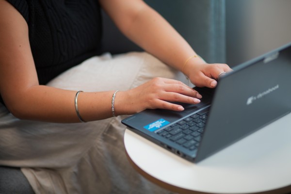 A woman (from check down) is sitting in a chair typing on her black laptop.