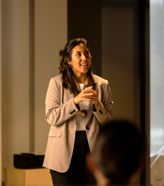A young woman with dark brown hair and a cream blazer looks off into the distance while delivering a presentation