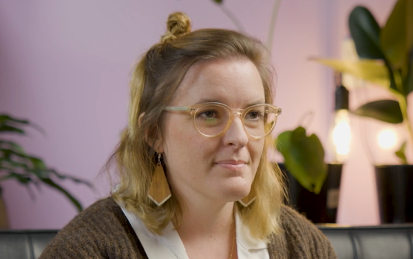 A woman sits inside on a leather couch in front of a purple lit wall and indoor plants. She has blond hair pulled into a bun and is wearing glasses.