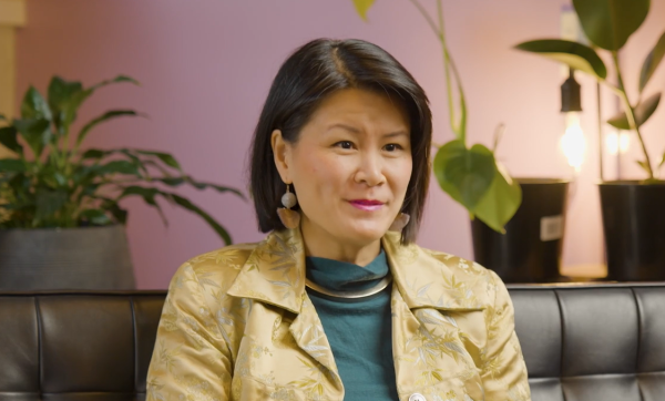 A woman sits inside on a leather couch in front of a purple lit wall and indoor plants. She has short brown hair and is wearing a yellow jacket.