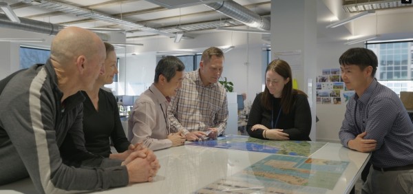 Six people standing around a table in an office studying a map